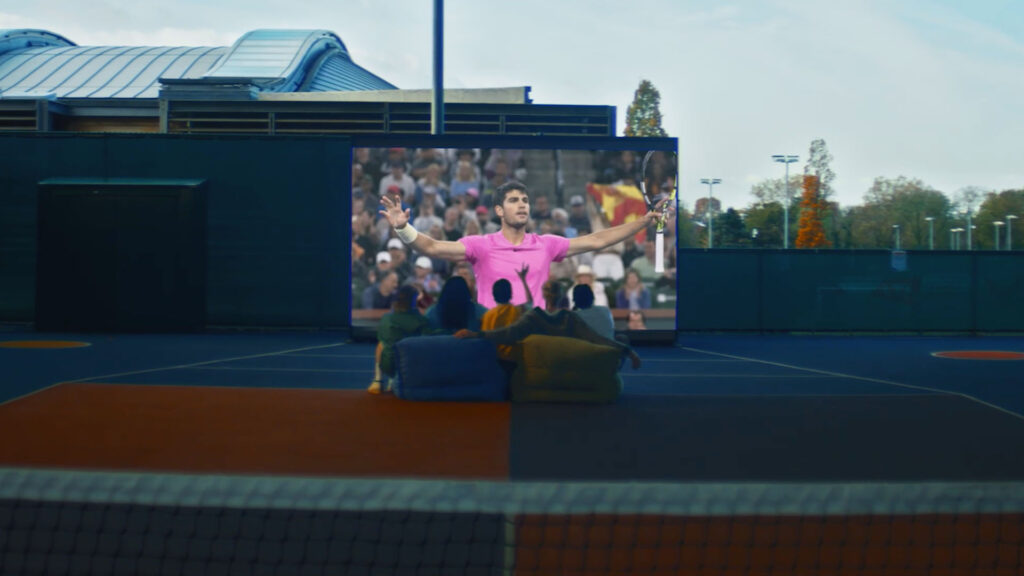 People sitting on a tennis court watching Carlos Alcaraz on a large screen