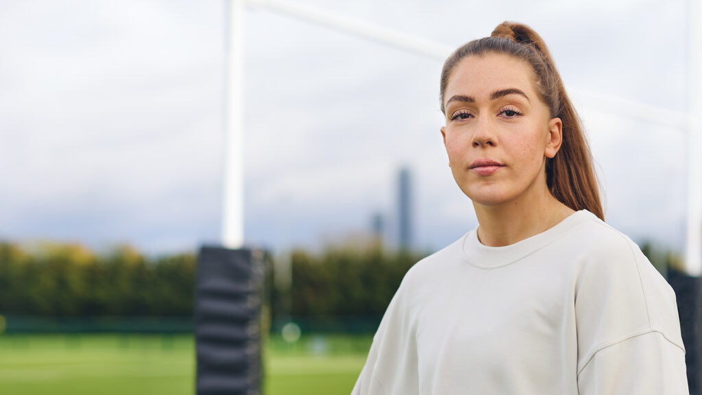 Holly Aitchison image in front of rugby goal posts in a white jumper