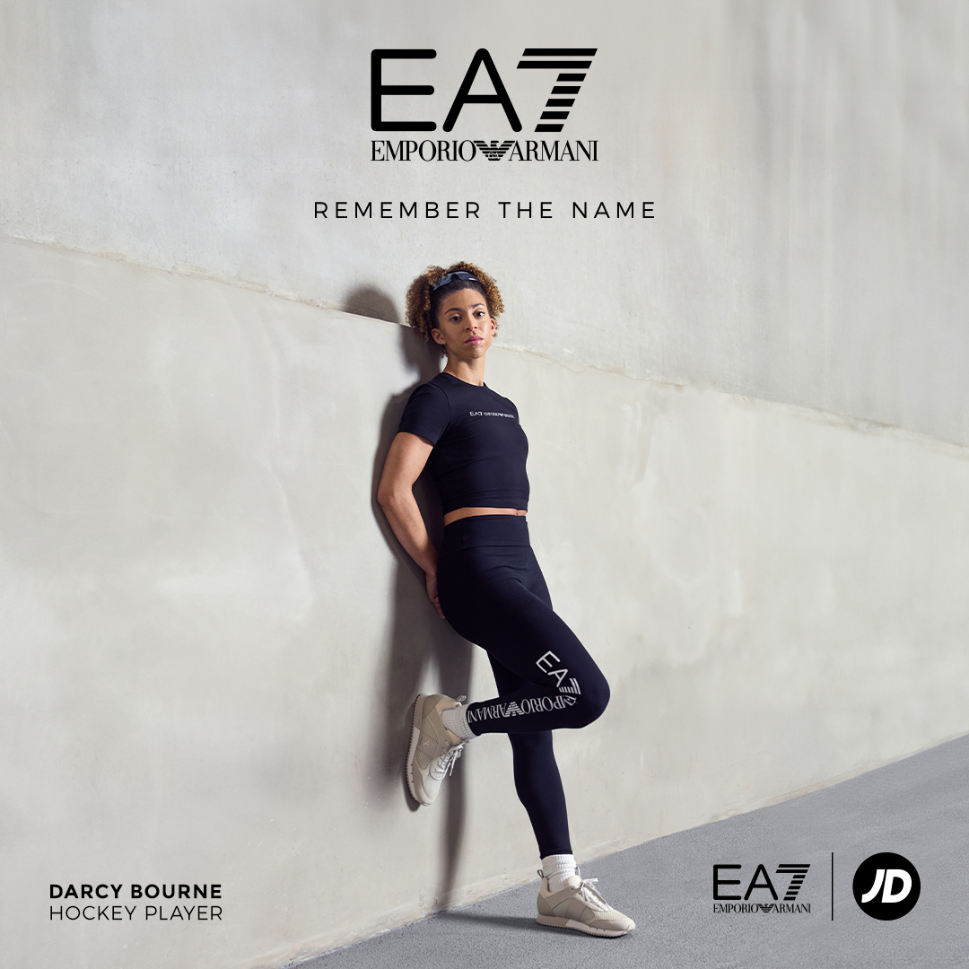A young female athlete leans against an industrial off-whiet wall wearing EA7 activewear, with the EA7 logo overlaid along with the headline REMEMBER THE NAME