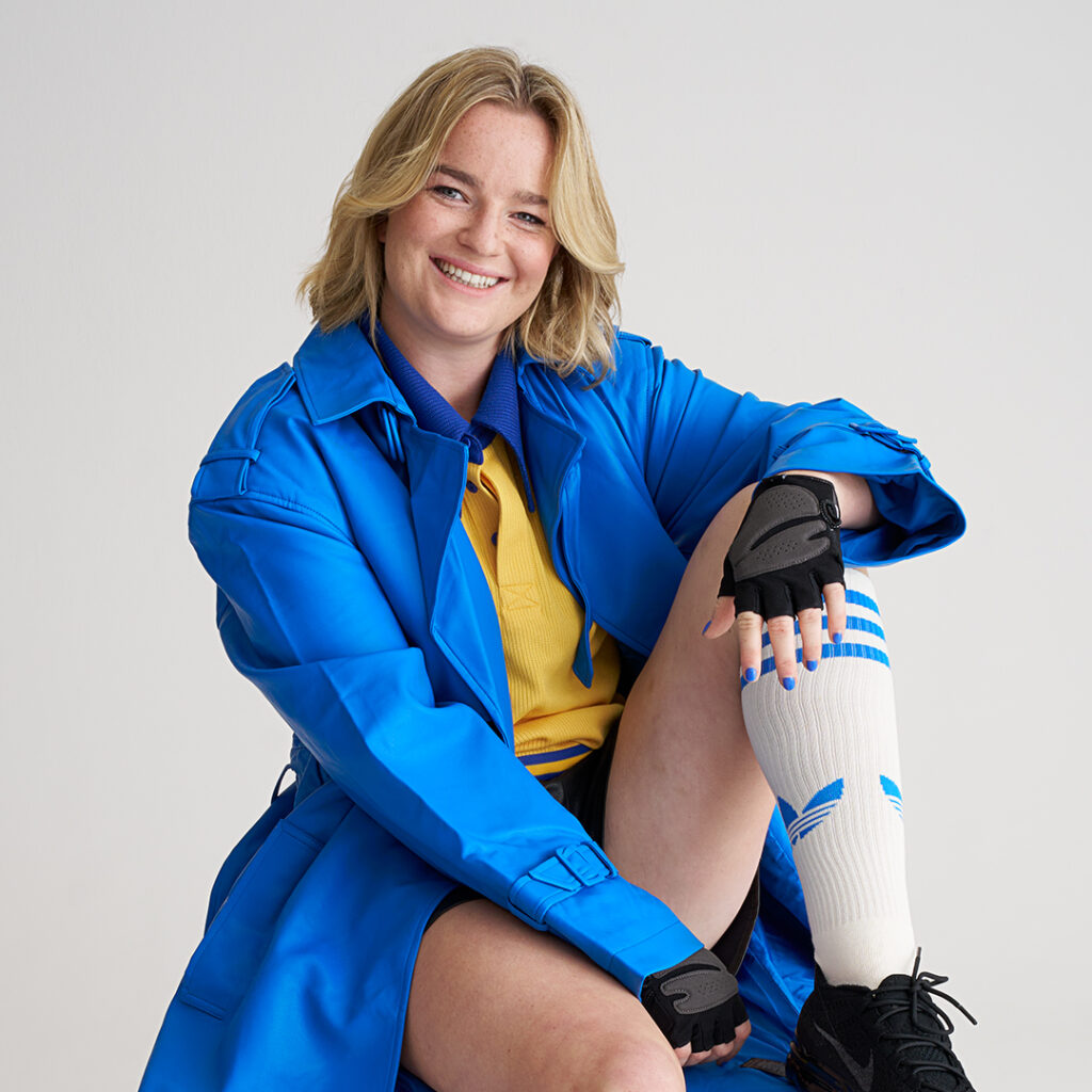 Daisy Hibert-Jones sat smiling in front of a white backdrop with a blue coat and yellow t-shirt on