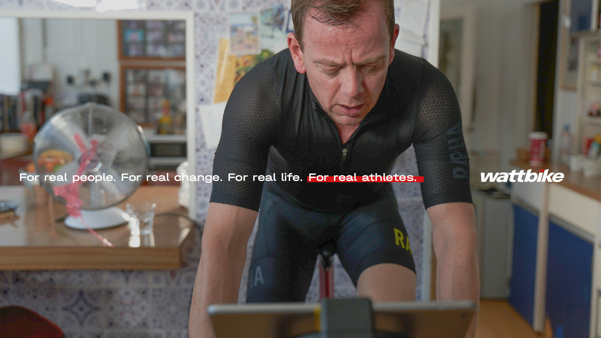 A man sitting on a Wattbike in his kitchen looking tired from his workout. A title on screen reads: “For real people. For real change. For real life. For real athletes.”, alongside the Wattbike logo.