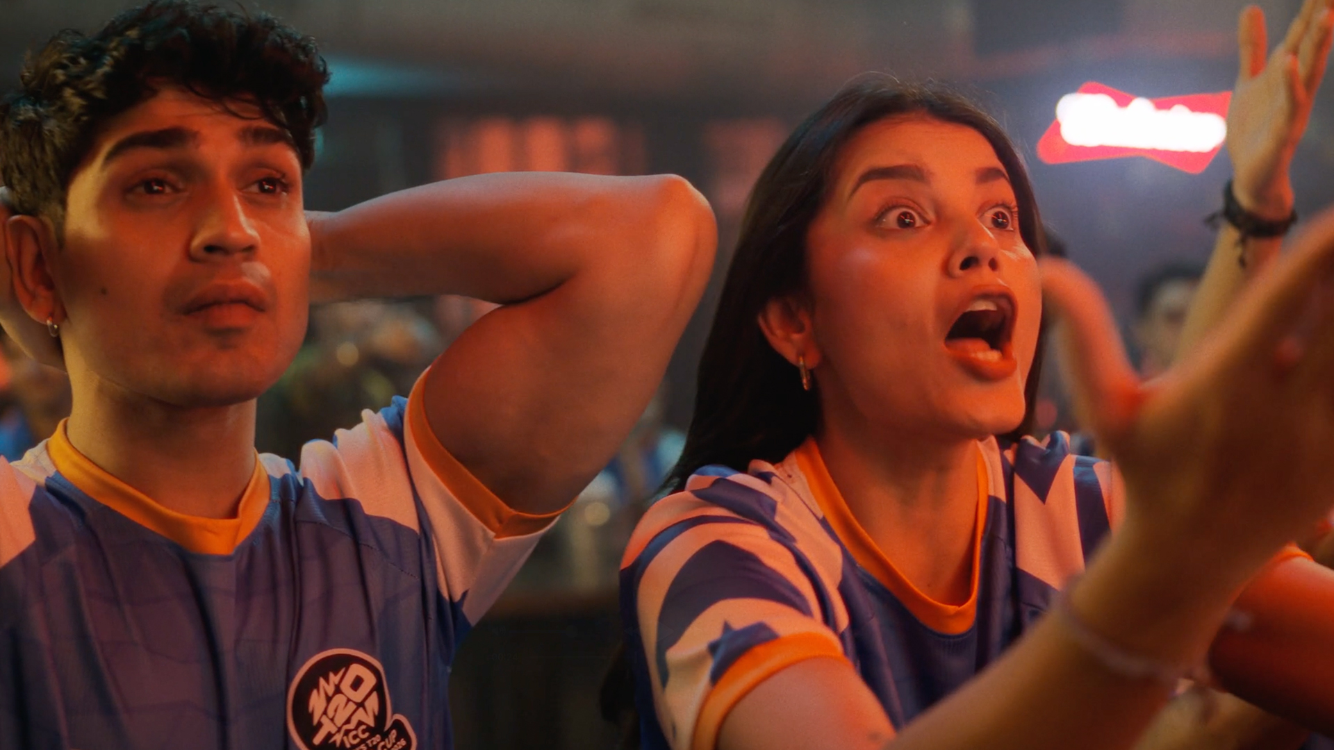 Two cricket fans wearing blue T20 jerseys react emotionally in a crowded indoor venue, watching a match with shock and excitement