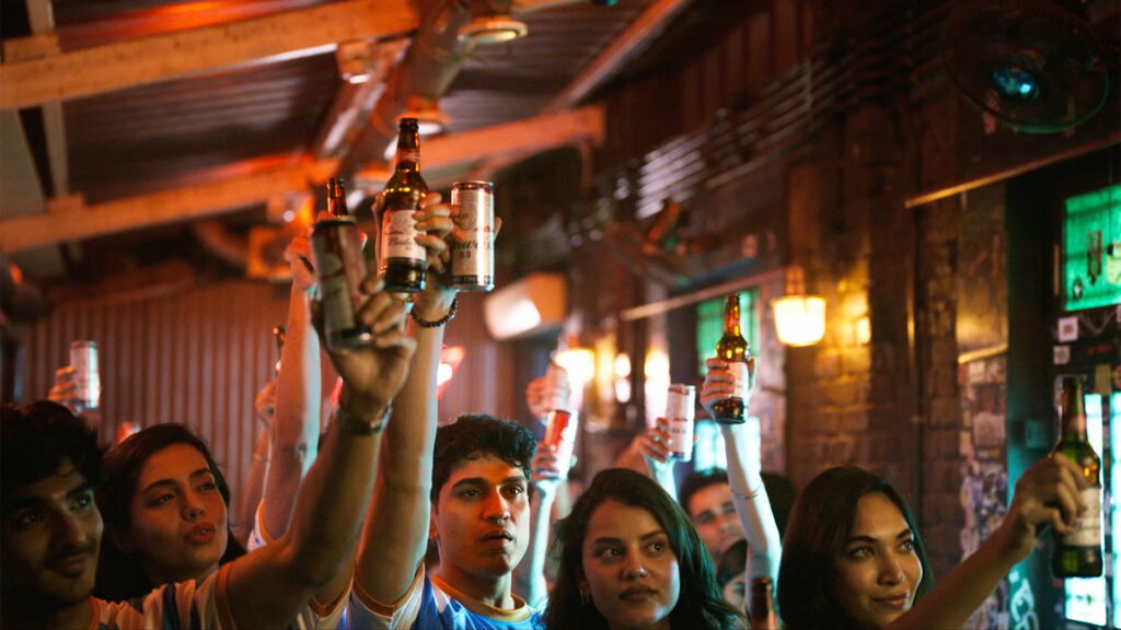 A group of cricket fans in a crowded indoor venue raise Budweiser bottles and cans in the air together