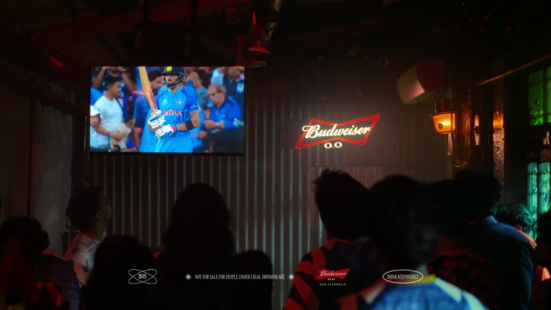 A crowd of cricket fans stand watching a live match on a wall-mounted screen in an indoor venue, with Budweiser 0.0 branding illuminated beside it
